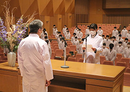 医療系大学の講堂で、白衣を着た学生が壇上から演説を行い、他の学生たちが真剣に聞き入っている。