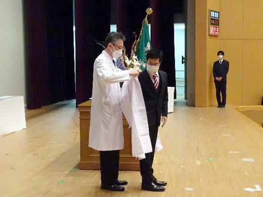 白衣を着た医師が、壇上で学生に白衣を着せている場面。後方にはスーツ姿のスタッフ。