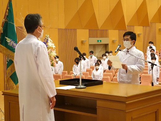 医学生が式典で宣誓し、壇上に立つ教授と対面。背後に多くの白衣を着た学生たちが見守っている。