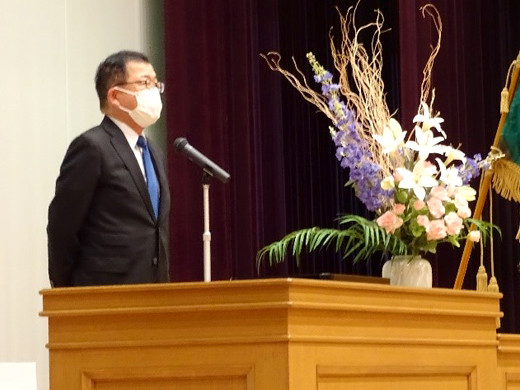 壇上でスーツを着たマスク姿の男性がマイクの前に立ち、隣に花瓶に活けられた色とりどりの花が配置されています。