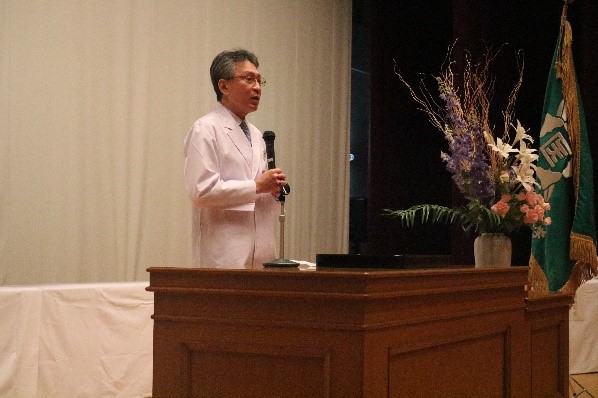 白衣を着た男性が壇上でマイクを持ち講演しています。背景には花の装飾と大学の旗があります。