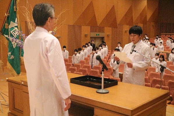 講堂内で医学生が壇上で宣誓を行い、聴衆は白衣を着用し整列している場面。