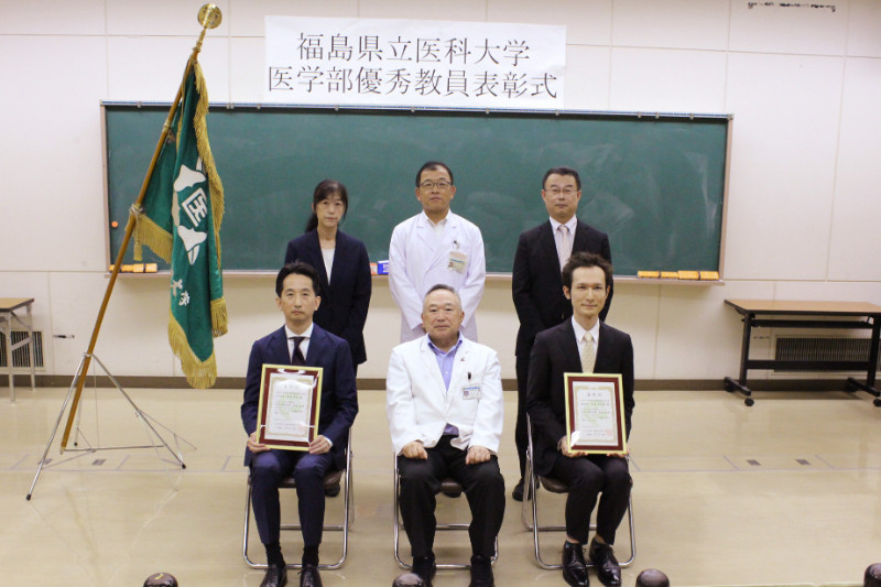 福島県立医科大学の優秀教員表彰式で、前方に証書を持つスーツ姿の男性2名と白衣の男性、背後に3名の立っている人物がいる。