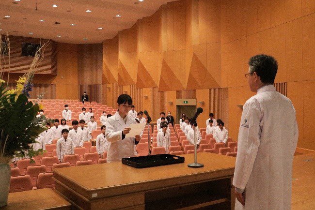 講堂で白衣を着た医学生たちが前方に立つ医師から指導を受けている様子。背後の座席には学生が並ぶ。