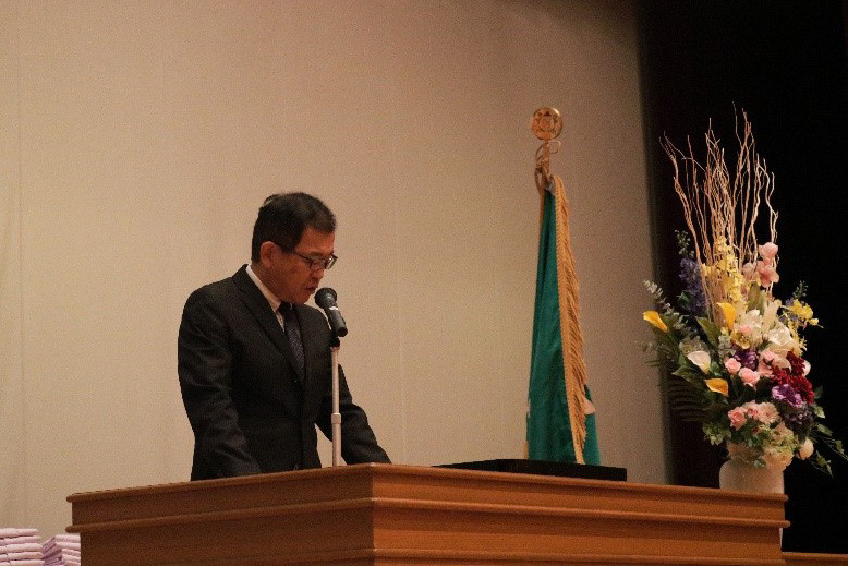 壇上でスーツを着た男性がマイクの前で話している。背景に学旗と花が飾られている。