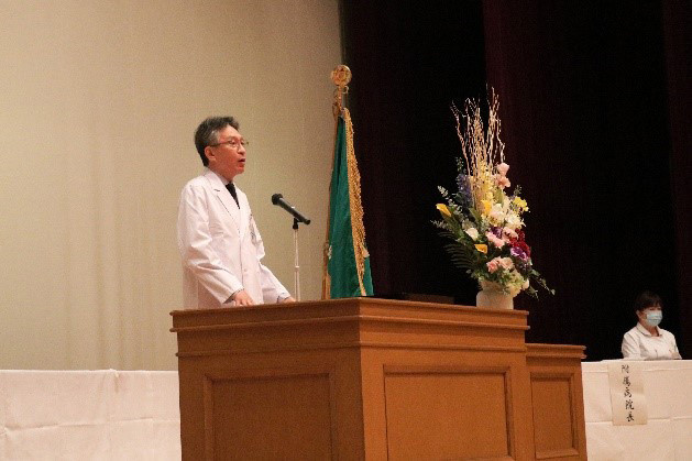 白衣を着た男性が講台に立ち、壇上でスピーチをしています。背景には大学の旗と花が飾られています。