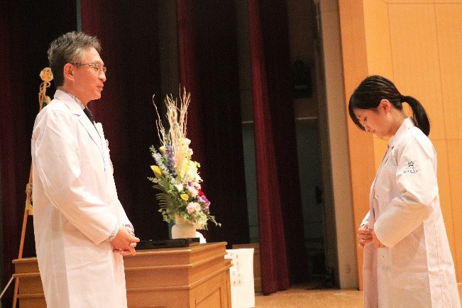 白衣を着た男性が講台に立ち、壇上でスピーチをしています。背景には大学の旗と花が飾られています。