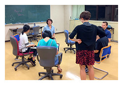 教室で円になって座っている5人の学生たち。黒板が背景にあり、カジュアルな服装で対話中。