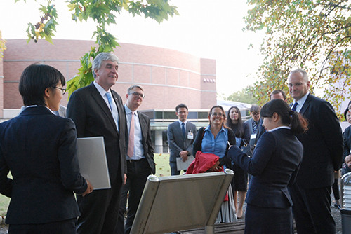 グループのビジネススーツ姿の大人たちが、屋外で看板の前に集まり話しています。背景にレンガの建物と木々があります。