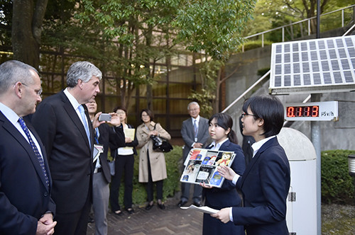 グループのビジネススーツ姿の大人たちが、屋外で看板の前に集まり話しています。背景にレンガの建物と木々があります。