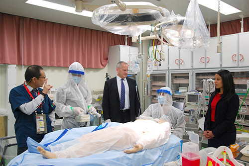 医療シミュレーション中の医療スタッフとマネキン。防護服を着た医療従事者とビジネススーツの観察者が周囲に立っている。