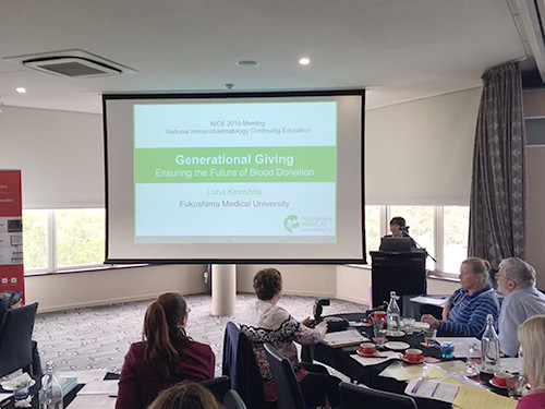 会議室でスライドを使ったプレゼンテーションが行われており、参加者たちが真剣に耳を傾けている。