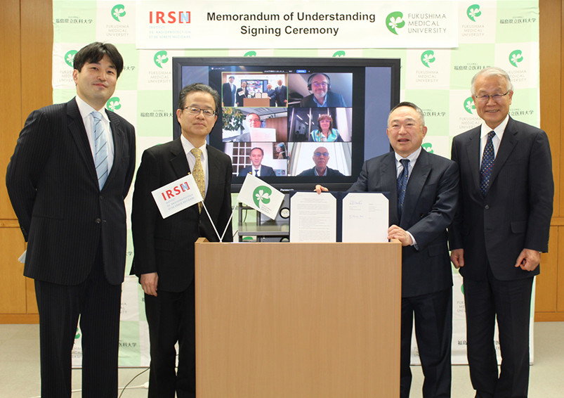 5人の男性が署名式で一緒に立つ。背後にビデオ会議の画面があり、複数の参加者が映っている。テーブル上に契約書が置かれている。