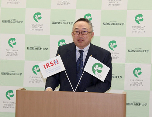 福島県立医科大学の背景を背にした講演台で、スーツ姿の男性が講演を行っている。