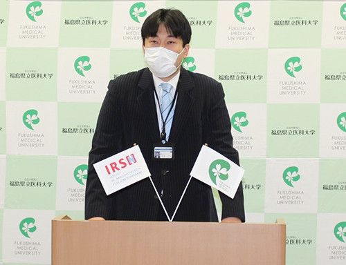 スーツ姿の男性がマスクを着用し、講演台の後ろに立っている。背景には「福島県立医科大学」のロゴが繰り返し配置されている。