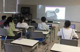 教室で数名の学生がプロジェクターのスライドを見ながら授業を受けています。前方のホワイトボードに学習内容が映し出されています。