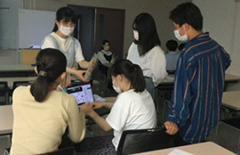 教室で数人の学生がテーブルを囲み、ノートパソコンを使ってディスカッションしています。全員がマスクを着用しています。