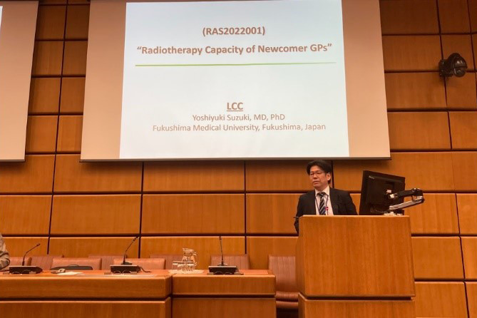 医科大学の教授が講演し、スクリーンに「Radiotherapy Capacity of Newcomer GPs」のスライドが映っている会議室の様子。