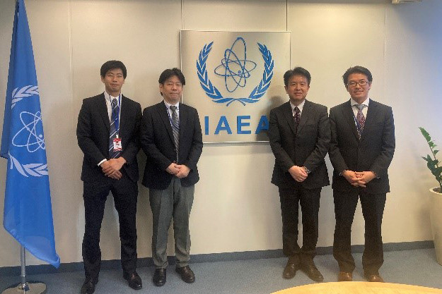 4人の男性がIAEAのロゴがある壁の前にスーツを着て並んで立っている。左には国連の旗が立てられている。