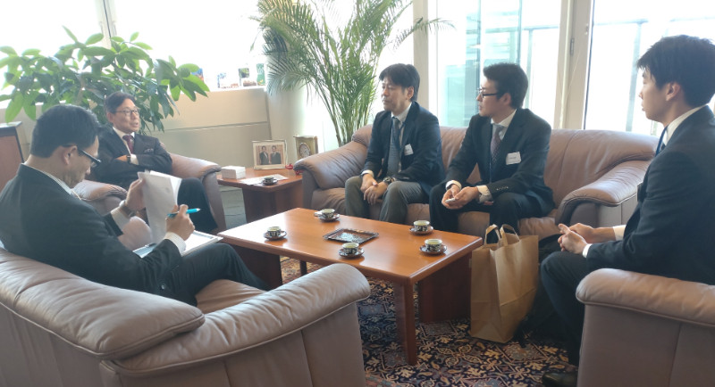 ソファに座ったスーツ姿の5人の男性が、コーヒーカップを前にテーブルを囲み会話している。背景には観葉植物と写真が飾られている。