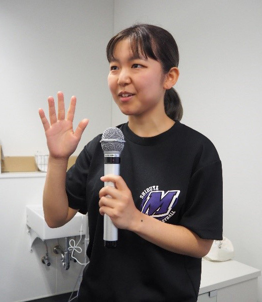 マイクを持ち、手を挙げて話す黒いTシャツの若い女性。室内にて講演やプレゼンテーション中。