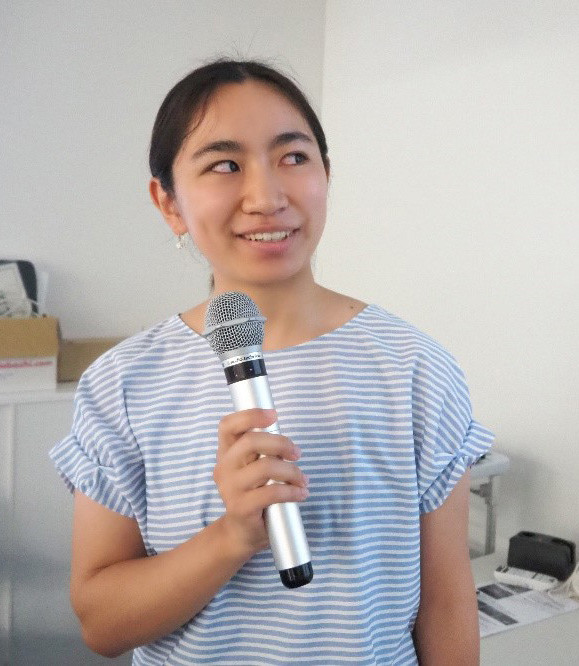 カジュアルな服装の女性がマイクを持ちながら話している様子。背景に白い壁がある室内。