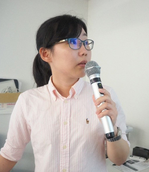 女性が教室でマイクを持って話している様子。彼女は眼鏡をかけ、カジュアルなシャツを着ている。背後に資料がある。