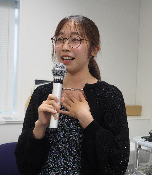 女性が教室でマイクを持ち、話している様子。彼女はカジュアルな服装で、真剣な表情を浮かべている。