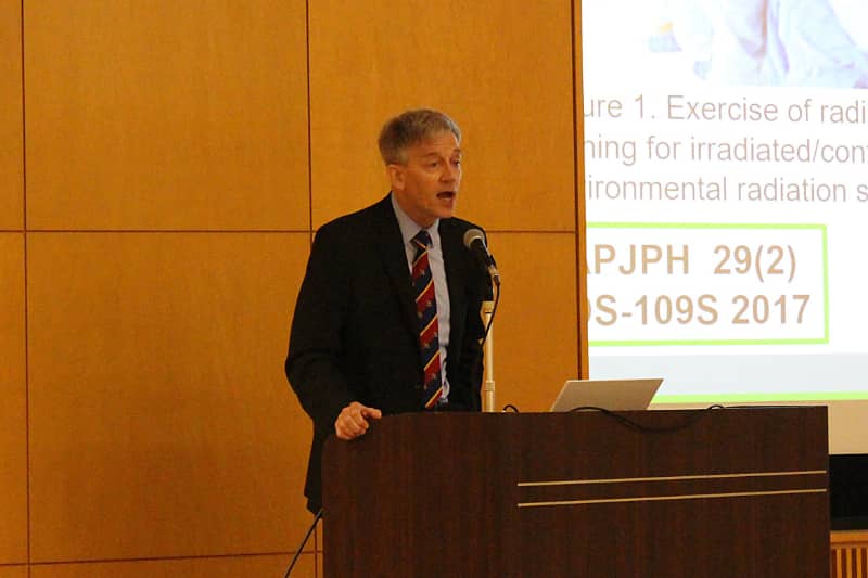 木製パネルの前で講演中の男性。スーツを着ており、プロジェクタースクリーンにはスライドが映し出されている。