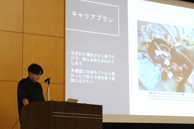 男性が講演台でプレゼンテーションを行い、スクリーンに「キャリアプラン」と題したスライドが投影されている。スライドには、医療に関する課題と彼の志望についてのテキストが表示されている。