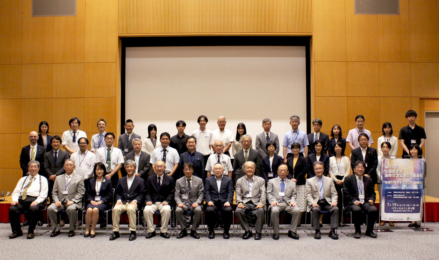 多くの大学関係者がホール内で二列に並び集合写真を撮影しています。背景には大きなスクリーンと木の壁があります。