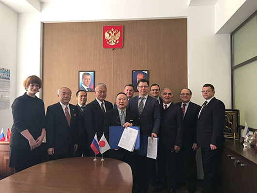 数名の男性と女性が会議室で協定書を持ちながら並んで立っており、日露の小さな国旗が机上に置かれています。