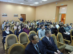 会議室で日本とロシアの旗の前に並ぶ複数の男性と女性。中心の二人は書類を持って微笑んでいる。背景にはロシアの紋章と写真が飾られている。