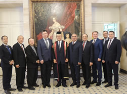 複数のスーツ姿の男性たちが美術品が飾られた室内で並んでいる。中央の人物はガウンと帽子を着用している。