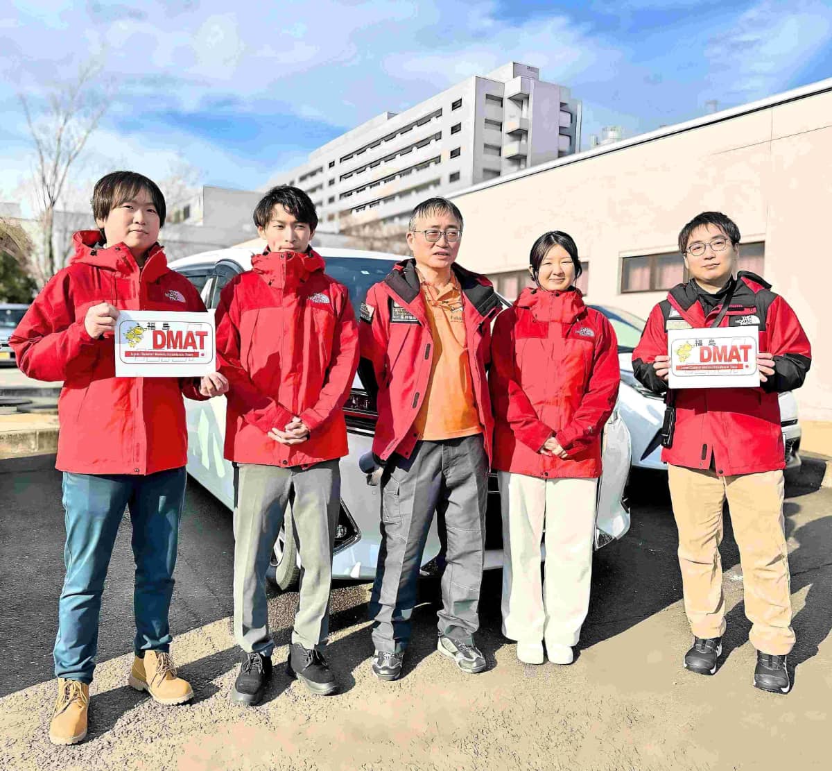 赤いジャケットを着た医療チームが屋外で並んで写真に写っており、「DMAT」と書かれたカードを持っている。背景に建物と車が見える。