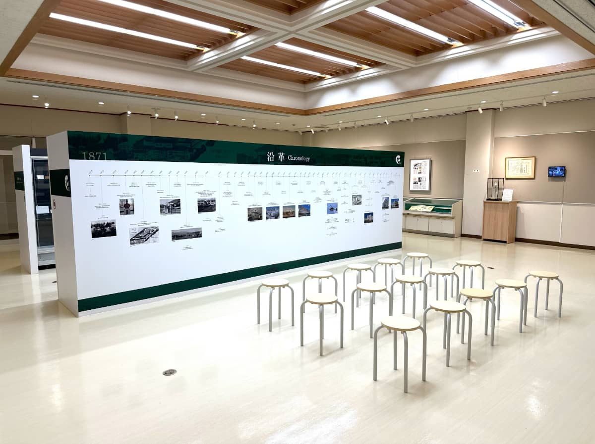 展示会場の様子。壁には大学の歴史を示す年表があり、その前に幾つかの椅子が並んでいる。天井には照明がある。