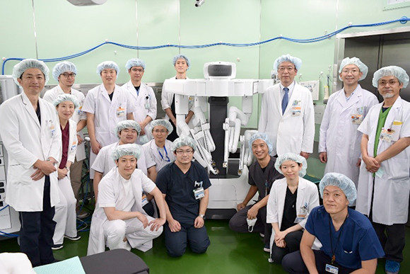 手術室で手術用ロボットを囲む医師たちのグループ写真。全員が手術帽を着用し、明るい緑の背景が特徴。