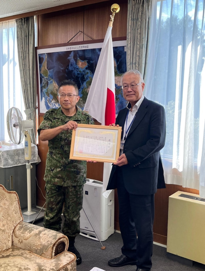日本の国旗の前で、軍服の男性とスーツ姿の男性が賞状を持っているオフィス内の様子。