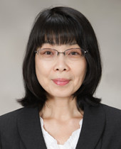 女性が黒のスーツを着てカメラに向かって微笑んでいる。背景は単色で、彼女はメガネをかけている。