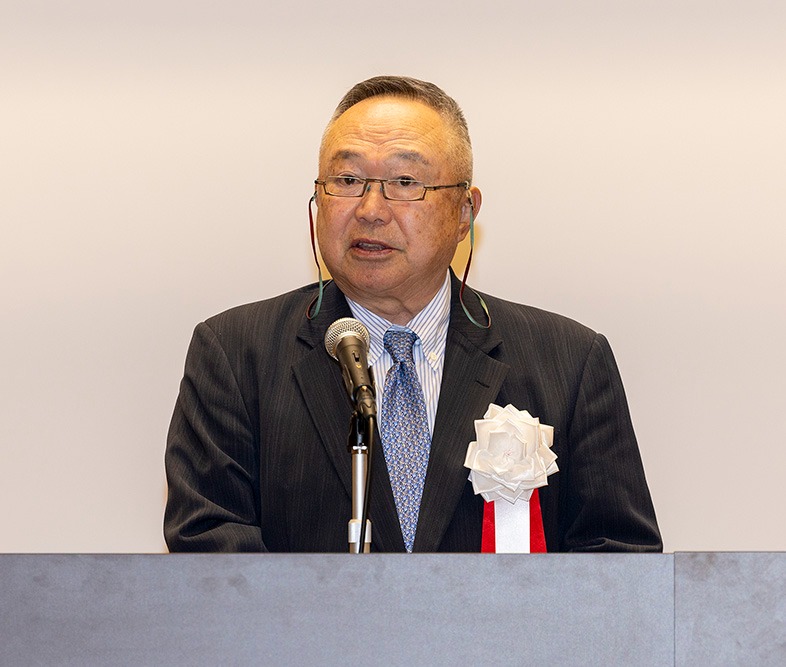 シニアの男性がスーツ姿でマイクの前に立ち、スピーチを行う様子。胸には白い花の装飾が付いている。