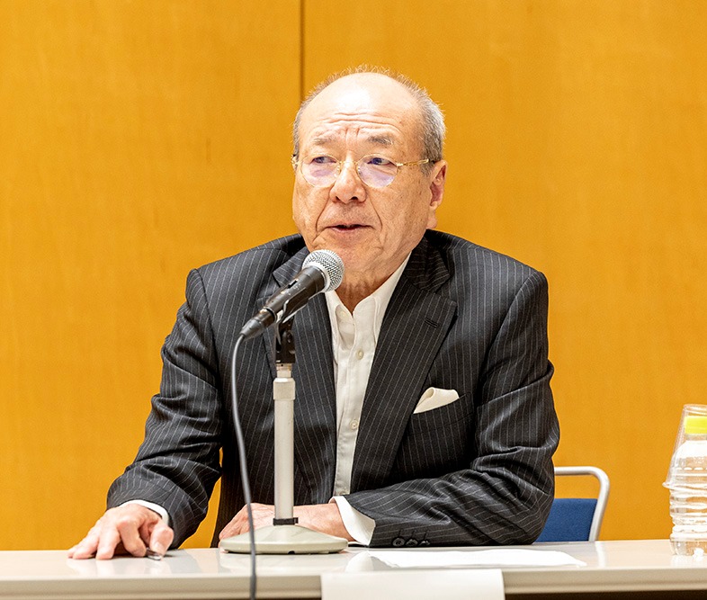 年配の男性がスーツを着てマイクの前に座り、会議または発表の場で話している。