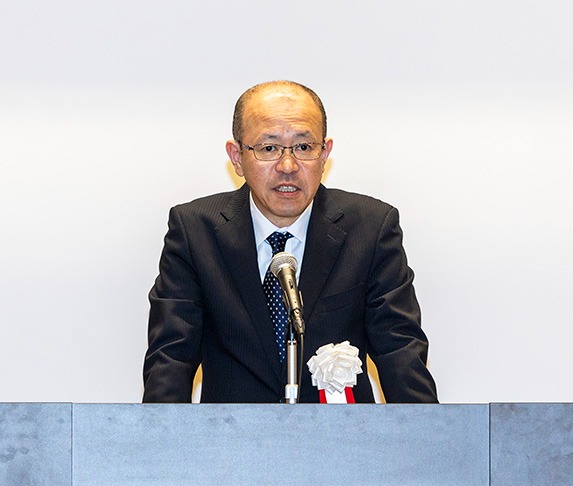 壇上でスーツを着た男性がマイクの前で講演している。白い花の飾りが胸に付いている。背景はシンプルな白。