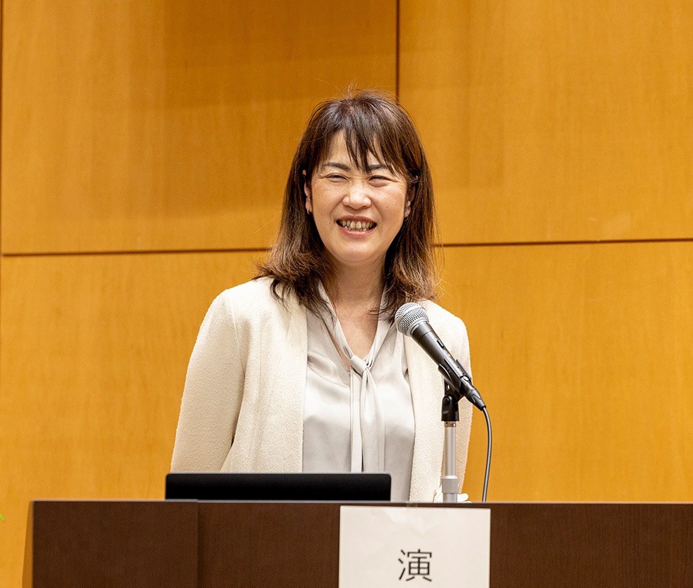 壇上でマイクを持ちながら笑顔で講演をしている女性。背景は木目調の壁。