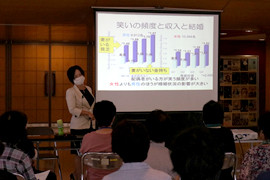健康の効果について講演する女性講師と、参加者が集まる会場の様子。