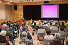 健康長寿をテーマにした講演会で、講師がスライドを使って説明している様子。聴衆は高齢者が中心。