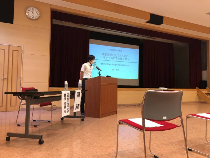 健康寿命を延ばすための講演会で、講師がマイクの前に立ち、スライドを使って説明している様子。