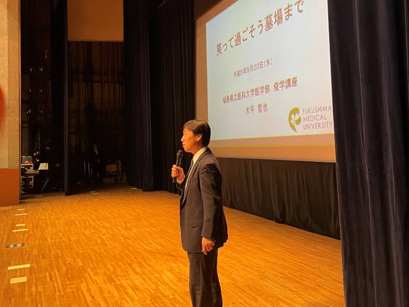 講演会で話す大平哲也教授、テーマは「笑って過ごそう墓場まで」。背景にはスライドが映し出されている。