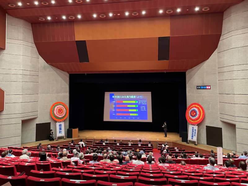 講演会の会場で、スライドを使って「笑いと健康」について説明する講師と聴衆の様子。