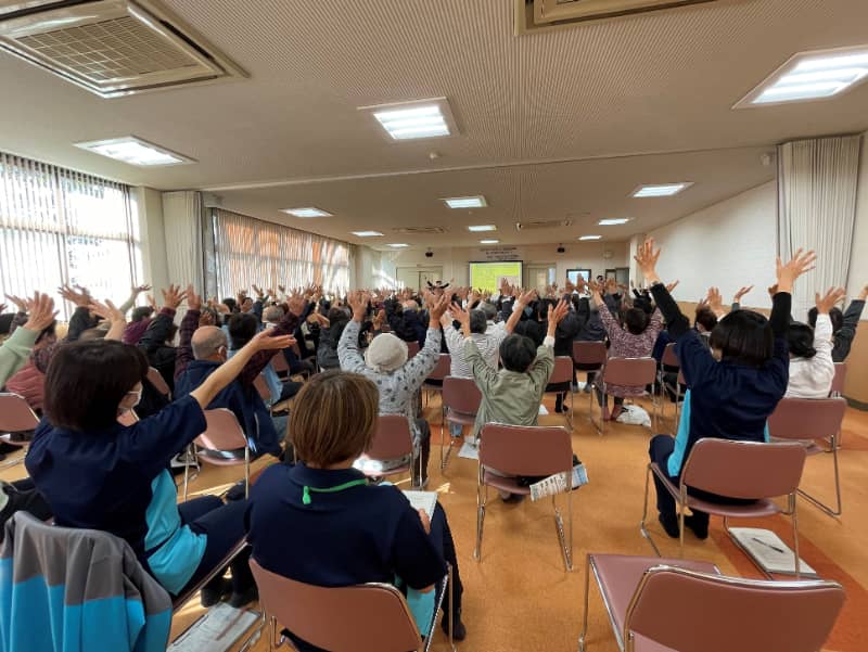 参加者が椅子に座り、手を挙げて笑いヨガを実践している様子。健康促進を目的とした活動の一環。