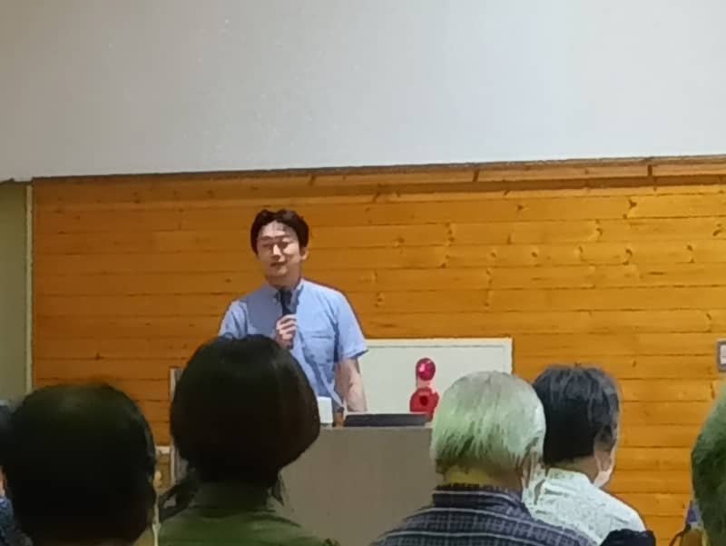 講演会で話す八巻尚洋准教授と聴衆の様子、背景には木の壁が見える。